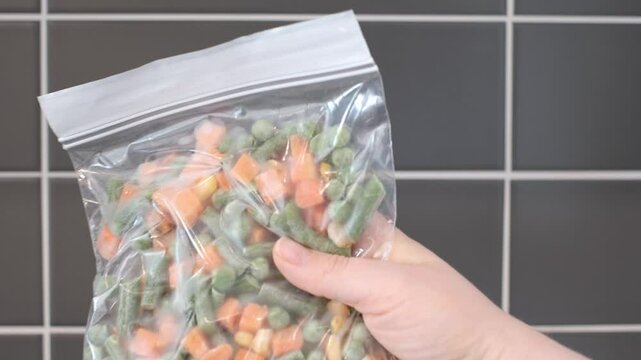 Female hand holding zip bag with a frozen chopped vegetables on gray tile background in kitchen.