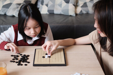 Fototapeta premium Asian family, mum, and kid fun playing Go, checkers board game on table in home living room together with love and cheerful, happy weekend relaxing hobby of child and parent, concentrate and compete.