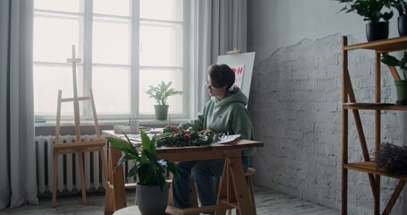 Young woman artist dressed in a hoodie draws pictures on paper with watercolors while sitting at a table in a workshop