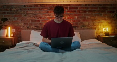 Young stylish man with tousled hair is using a laptop with a focused look, while sitting on the bed...