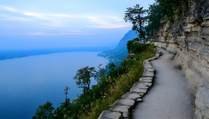 Obraz premium Scenic lakeside walking path carved into a cliffside, offering breathtaking panoramic views of serene blue waters and distant mountains at sunset.