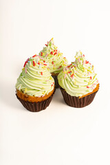  Three cupcakes with green frosting and colorful sprinkles sit on a white background. The frosting has a swirled texture, and each cupcake is in a dark brown paper cup.