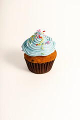 A cupcake with blue frosting and colorful sprinkles sits on a white background. The frosting has a swirled texture, and the base is in a brown paper cup