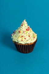 A cupcake with green frosting and colorful sprinkles sits on a blue background. The frosting has a swirled texture, and the base is in a dark brown paper cup.