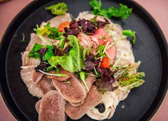 A variety of meat snacks, boiled beef tongue, roast beef and meat roll on a black plate with lettuce leaves and microgreens. Restaurant dish on a light pink tablecloth
