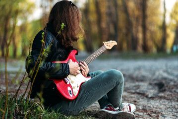 A guy with long hair plays an electric guitar on the riverbank, a non-binary person, a man plays a guitar in the forest, a guy in a red shirt and sneakers,a red guitar,a man learns to play the guitar