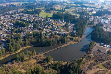Aerial drone view of a residential neighborhood in Tualatin, OR, featuring a small lake, tree-lined streets, modern suburban homes, a train track bridge crossing over the water in a peaceful setting