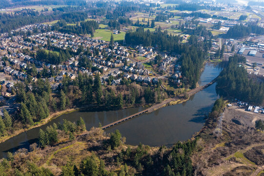 Aerial drone view of a residential neighborhood in Tualatin, OR, featuring a small lake, tree-lined streets, modern suburban homes, a train track bridge crossing over the water in a peaceful setting