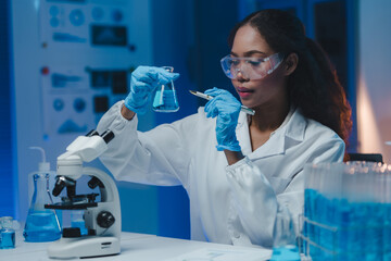 Female scientist wearing lab coat and gloves carefully examining beaker filled with blue liquid while holding pen in modern laboratory, microscope and test tubes visible in background