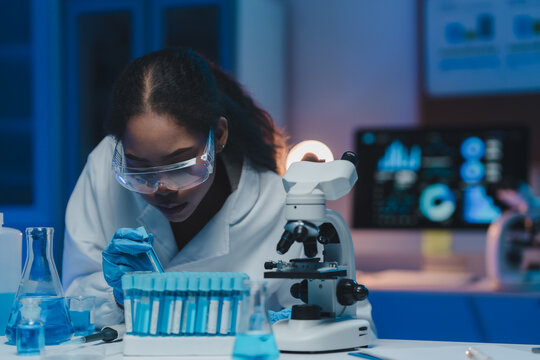 Young female scientist wearing lab coat and gloves carefully analyzing test tube with blue liquid, working with microscope and computer in modern laboratory at night - Powered by Adobe