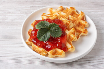 Delicious waffles topped with strawberry jam and a fresh leaf on a white plate with a light wooden background