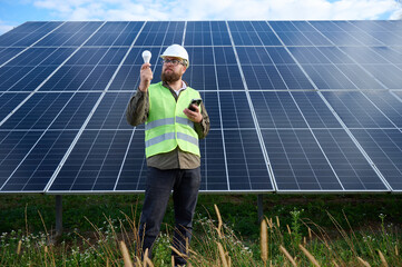 Engineer in uniform with protective helmet near solar station holding light bulb. Supervisor is...
