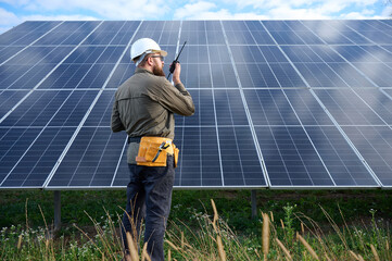 Engineer in uniform with protective helmet near solar station holding walkie talkie. Supervisor is...