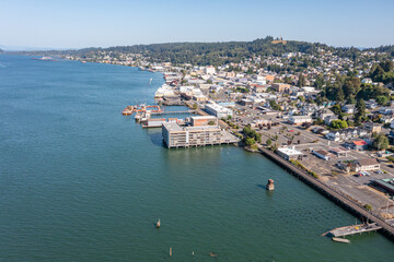 Aerial drone view of downtown Astoria, Oregon, featuring waterfront buildings, the Columbia River, a busy port, fishing boats, and lush hills in the background