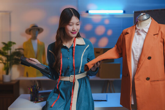 Young beautiful asian woman fashion designer holding digital tablet and checking quality of clothes on mannequin in modern studio, dressmaker working with online orders in atelier