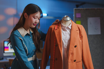 Young dressmaker adjusting measurements of an orange jacket placed on a dress form, working late at...