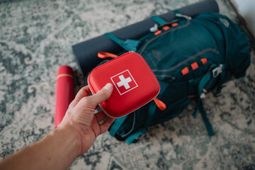 A person is holding a first aid kit in front of a backpack that seems ready for an outdoor...