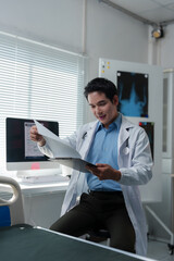 Young male asian doctor wearing lab coat and stethoscope reviewing patient medical records and test results on clipboard in hospital room with x-ray in background