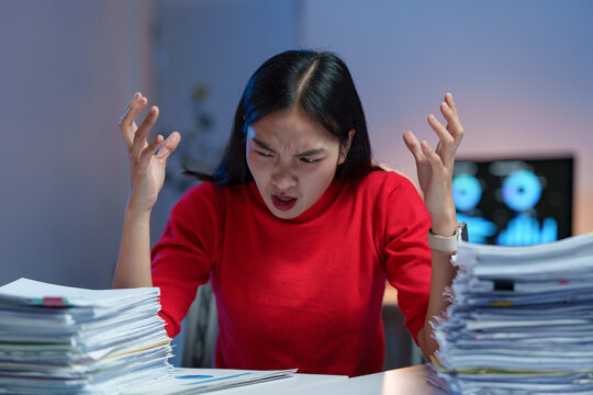 Young professional woman working late feeling stressed and frustrated by excessive paperwork, raising hands in despair while sitting at her desk in a dark office at night
