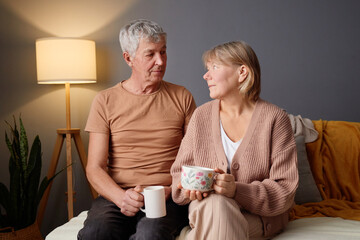 An elderly couple sits closely on a comfortable couch, sipping tea from decorative mugs. Soft light...