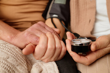 A healthcare worker measures blood pressure while holding hands with a patient in a cozy environment. The scene conveys support and trust during a medical examination