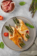 Baked puff pastry with green asparagus, prosciutto or ham and cheese on a gray concrete background. Top view.