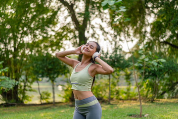 Young Asian woman enjoying music on headphones while exercising in green park. Sporty woman using...