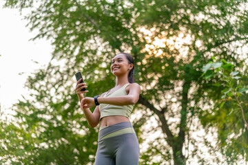 Young Asian Woman Using Phone and Smartwatch for Fitness Tracking in a Park. Beautiful Lady Embracing Health Technology in Outdoor Workout Routine
