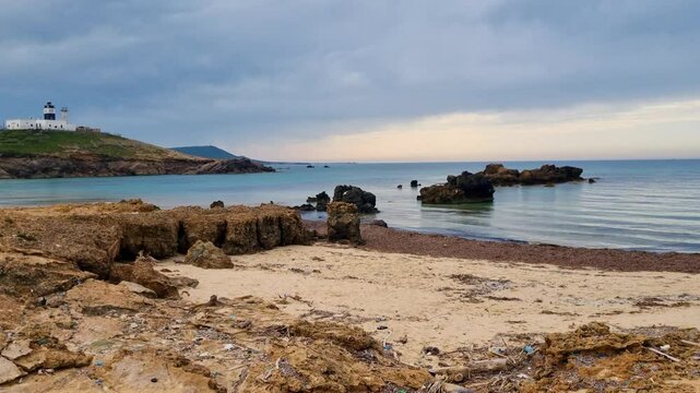 Panoramic shot of Cape Angela, the northernmost point of Africa, featuring the Mediterranean Sea, a deserted rocky beach, and a distant lighthouse perched on a green hill