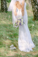 The bride in a white lace dress with a long veil holds a bouquet of flowers, outdoors, standing...