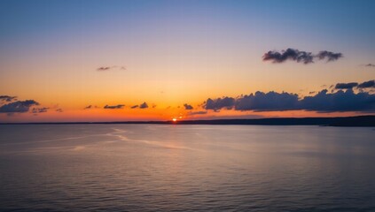 Fototapeta premium A serene sunset over a calm sea, vibrant orange and yellow hues blending into a soft blue sky, distant dark coastline, gentle waves reflecting the colourful light