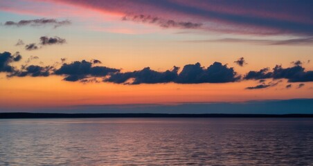 Obraz premium A serene sunset over a calm sea, vibrant orange and yellow hues blending into a soft blue sky, distant dark coastline, gentle waves reflecting the colourful light