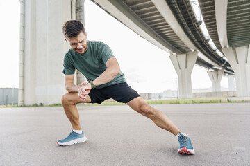 Man stretching under a bridge before jogging along a city pathway in the morning light