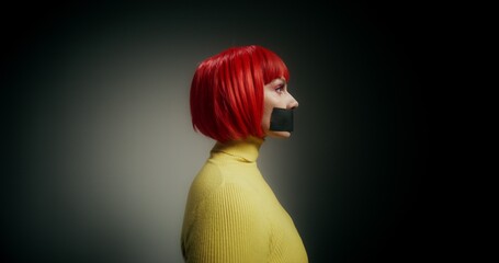 A woman in an orange wig with her mouth taped shut turns her head and looks at the camera with a...