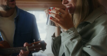 A man plays the ukulele and sings while looking at a woman sitting next to him on the bed. A woman drinks hot tea from a metal mug