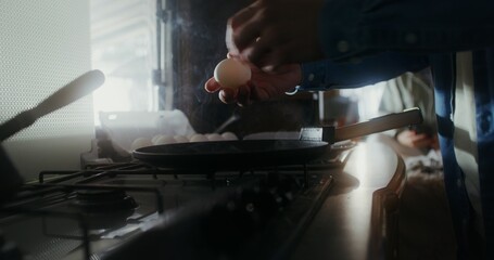Men's hands break eggs into a frying pan. A blurry woman is sitting on a bed in the background. Romantic journey in a van