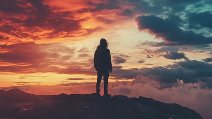 Obraz premium Silhouette of a woman standing on a mountaintop at sunset, with dramatic clouds overhead.