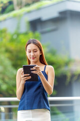 Fototapeta premium Young Asian Businesswoman In Casual Business Outfit Using Tablet Outdoors In Front a Modern Office Building