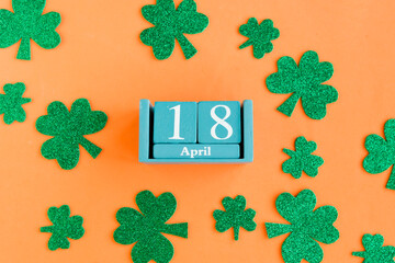 April 18. Blue cube calendar with month date and green clover leafs on bright orange background.