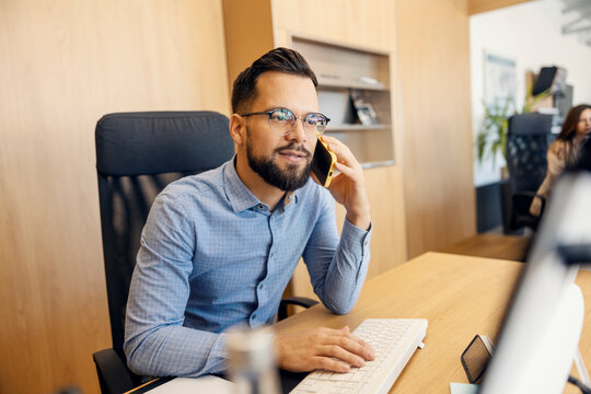 Portrait of a businessman sitting at his workstation at office in corporate firm with cellphone in hands and having phone call.