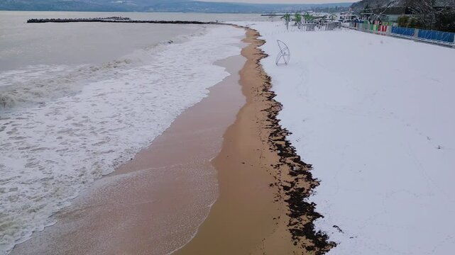 A unique winter beach scene where ocean waves meet the snow-covered shore. The contrast between sand, sea, and snow creates a stunning seasonal landscape with a peaceful and surreal atmosphere