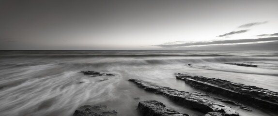 Black and white seascape with smooth waves crashing over rocky shoreline under cloudy sky at dusk