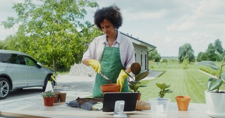Young woman in an apron and rubber gloves transplants flowers while standing in the backyard