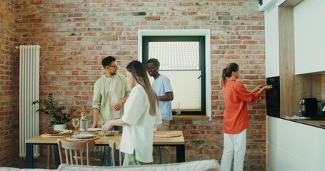 A group of young people sits down at a table in the kitchen, a woman takes a roast out of the oven,...