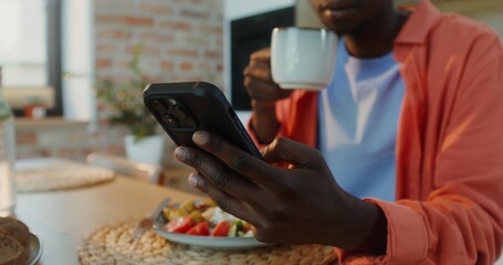 A man of African-American appearance uses a mobile phone while drinking coffee and dining at home, close-up of his hands, no face