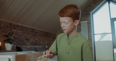 A red-haired boy paints a wooden shelf with a brush while sitting in a stylish home interior