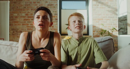 Mom and son of European appearance smile playing video games using joysticks sitting on the couch in a stylish home interior © m-art
