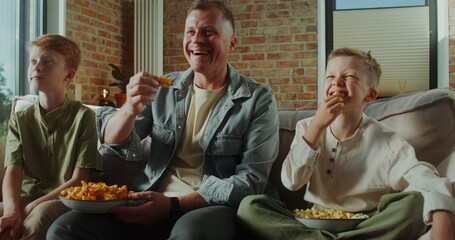 A man and two boys are sitting on the couch and eating snacks while watching TV at home