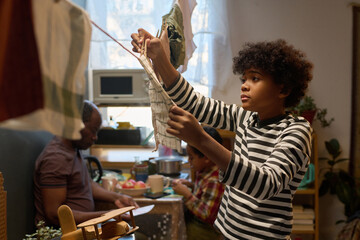 Young boy with Black person heritage hanging laundry in cozy home while sunlight filters through window, family members in background engaging in activities