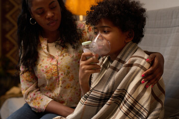 Mother comforting child using inhaler while sitting on sofa, wrapped in blanket and embracing him, young boy inhaling medicine with assistance from mother